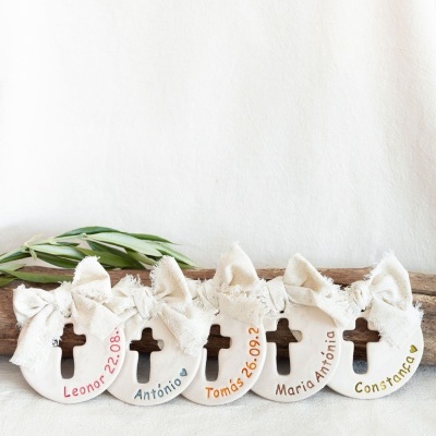 Cinco decorativos em cerâmica branca com nomes pintados e laços de tecido cru colocados em linha sobre fundo branco e tronco de madeira com folhas verdes ao redor.