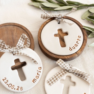 Adornos cerâmicos brancos com cruz recortada, nomes e laços xadrez sobre madeira e tecido.