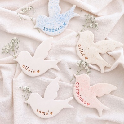 Cinco pássaros de cerâmica com nomes gravados e pequenos ramos de flores sobre tecido bege.