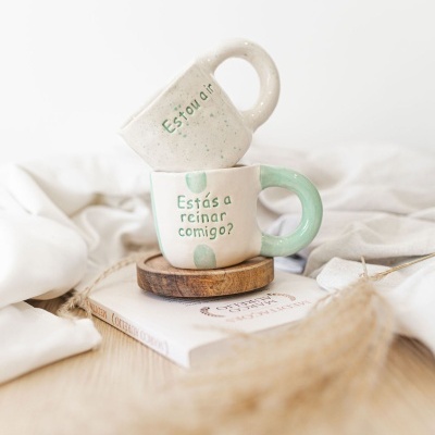 Duas chávenas de cerâmica branca e verde com texto em português, empilhadas sobre um prato de madeira e um livro