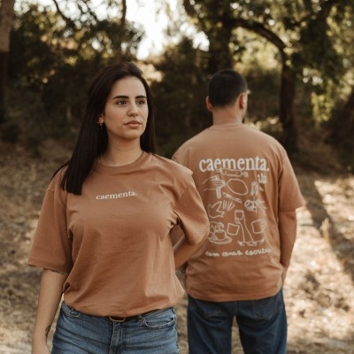 Duas pessoas com t-shirts castanho claro com texto branco caementa. e ilustrações, ao ar livre com árvores e luz do sol