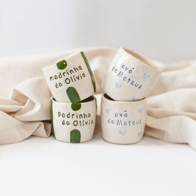 Conjunto de quatro canecas de cerâmica brancas com texto em verde e azul e manchas ou corações decorativos, em fundo claro soft.