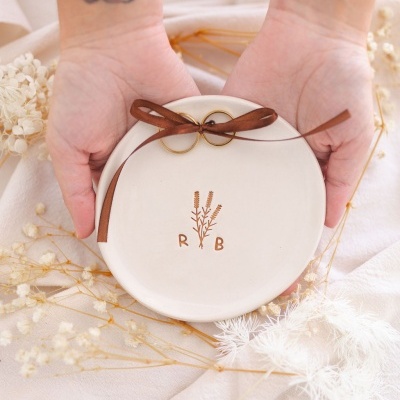 Prato cerâmico branco com alianças douradas amarradas com fita castanha, rodeado por flores secas e tecido claro.