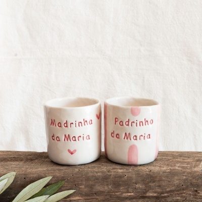 Dois copos de cerâmica artesanal com inscrições em português sobre um pedaço de madeira e fundo branco.