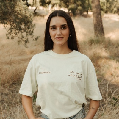 Mulher com t-shirt branca com texto bordado castanho e calças de ganga num ambiente campestre seco.