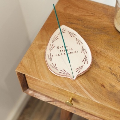 Suporte para incenso de cerâmica branca com padrão de ramos e texto 'Então repare na bagunça!' sobre mesa de madeira.