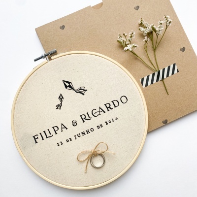 Argola bordada em tecido com nomes FILIPA & RICARDO, data 23 DE JUNHO DE 2024 e decoração de pipas, junto a envelope castanho com corações e flores secas