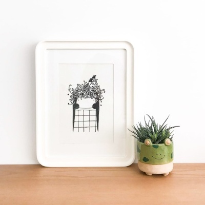 Moldura branca com ilustração de cadeira e plantas ao lado de vaso de planta decorativo tipo rã