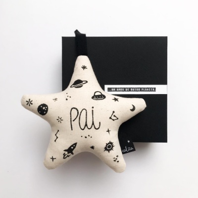 Almofada estrela creme com desenhos espaciais e a palavra 'Pai', sobre fundo branco e caixa preta com texto.