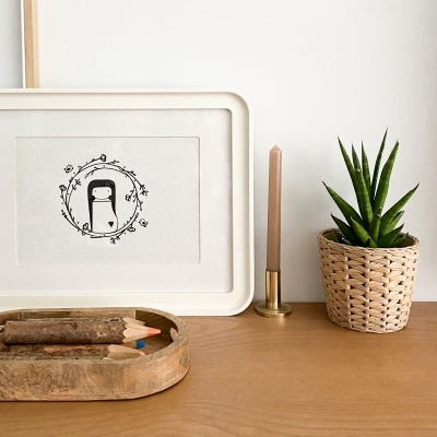 Mesa com lápis de cor grandes, moldura branca com desenho minimalista, castiçal dourado com vela e vaso de planta em cesto de palha.