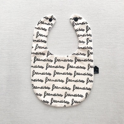 Babete branco com padrão repetido da palavra FRANCISCO em letras pretas sobre fundo cinzento claro.