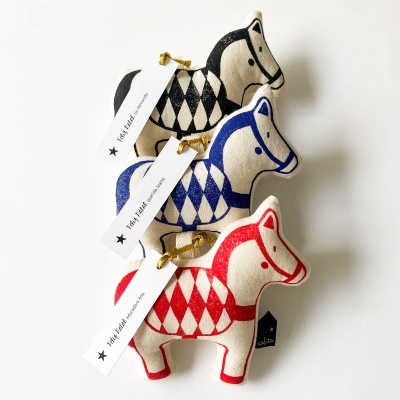 Três bonecos de tecido com padrão de losangos em preto, azul e vermelho, com etiquetas brancas e preta sobre fundo branco