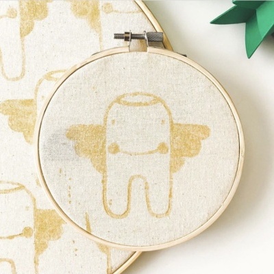 Aro de bordado em tecido bege com personagem dourado com asas.