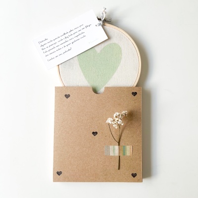 Quadro redondo com coração verde em tecido e envelope decorativo com flor seca e etiqueta com texto em português.