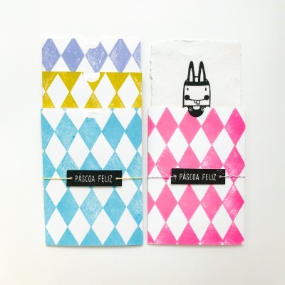 Caixas coloridas com padrão de losangos e texto PÁSCOA FELIZ, desenho de coelho visível num papel dentro de uma delas