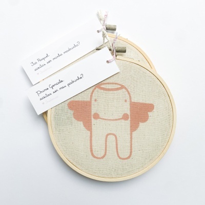 Dois bastidores de bordado em madeira com desenho rosa em tecido bege e etiquetas com texto em português.