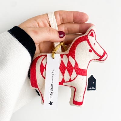 Decoração pendente em forma de cavalo branco com padrões vermelhos, feita em tecido, segurada por uma mão