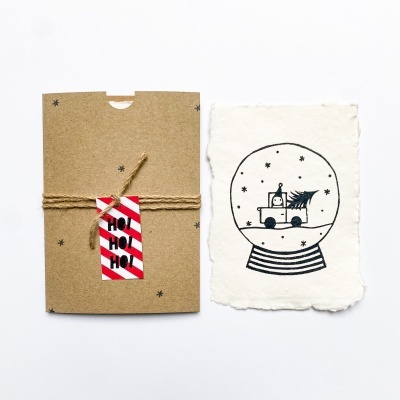 Pacote de cartão com etiqueta 'HO! HO! HO!' e cartão com desenho de globo de neve com veículo e árvore de Natal