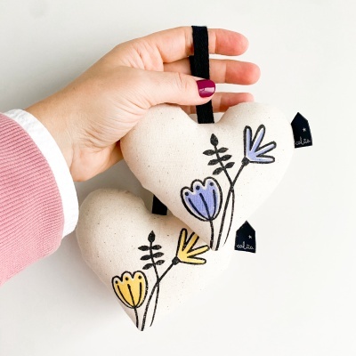 Dois corações de tecido com flores bordadas e etiquetas pretas, segurados por uma mão.