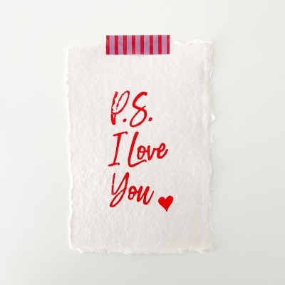 Papel artesanal branco com frase em vermelho P.S. I Love You preso com fita adesiva vermelha e roxa às riscas