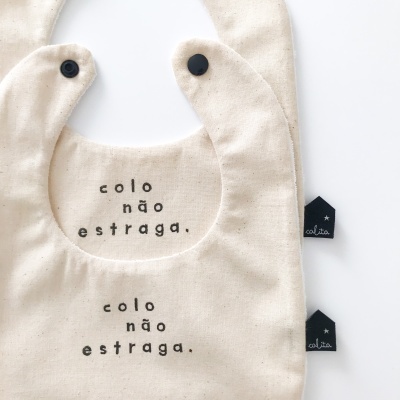Dois babetes de tecido cru com texto preto 'colo não estraga.' e fechos de pressão