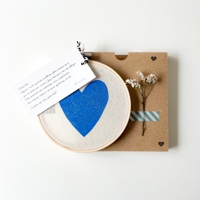 Bastidor de madeira com tecido branco e coração azul, cartão com texto, flores secas e caixa de cartão castanha.