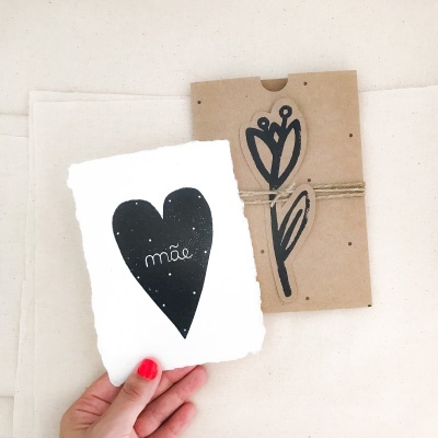 Cartão branco com coração preto e texto 'mãe', embalagem de cartão castanho com desenho de flor e cordel