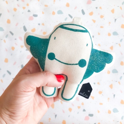 Pelúcia branca e verde-azulada com asa e sorriso segurada por mão com unhas vermelhas