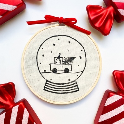 Aro de bordado com desenho de carrinha e árvore de Natal, rodeado de decorações natalícias vermelhas