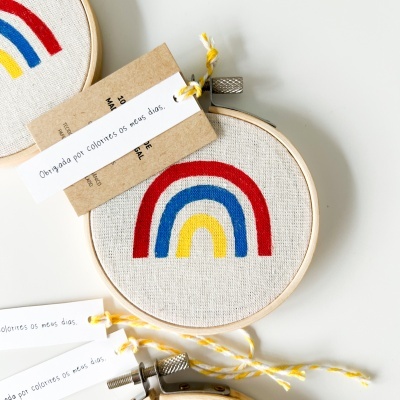 Bastidores de bordado com arco-íris colorido e etiquetas com texto em cordão amarelo e branco.