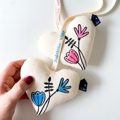 Dois corações de tecido branco com flores azuis e cor-de-rosa, etiqueta azul, fita colorida e mão com unhas roxas segurando um coração.