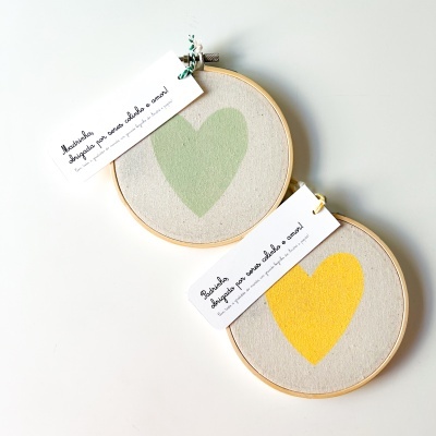 Dois bastidores de madeira com tecido bege e corações verde e amarelo, com etiquetas brancas com texto em português.