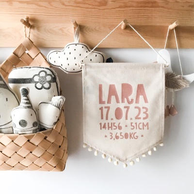 Placa decorativa de tecido com informações de nascimento pendurada na parede com cesto de vime e almofadas decorativas ao lado