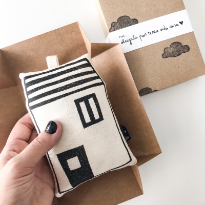 Almofada em tecido branca com desenho de casa a preto, mão a segurá-la e embalagem castanha com etiqueta branca e texto em português.