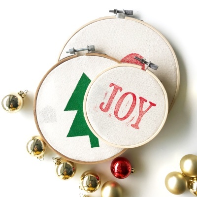 Três bastidores de bordado com motivos natalícios e bolas de Natal ao redor.