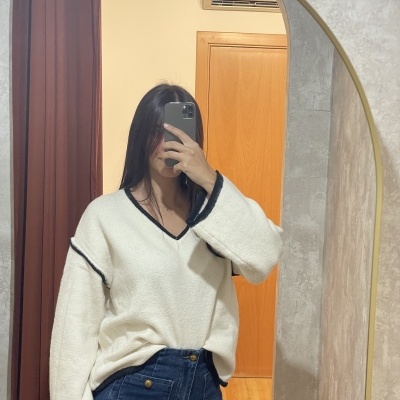 Mulher a vestir suéter branco com bordas pretas e jeans azul escuro refletida num espelho com moldura dourada.