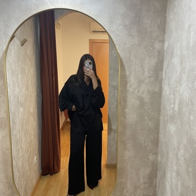 Espelho oval dourado numa parede cinza com reflexo de pessoa vestindo roupa preta e sapatos vermelhos