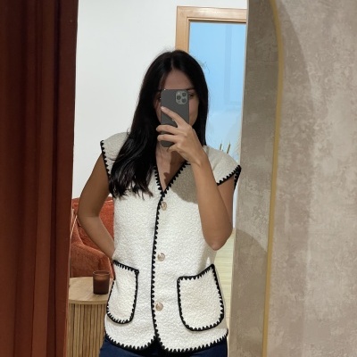 Colete feminino branco peludo com bordas pretas e botões grandes, com dois bolsos frontais, refletido em espelho