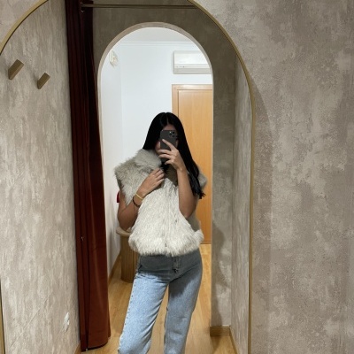 Espelho dourado em arco numa parede cinza, refletindo pessoa com colete de pele e calças de ganga