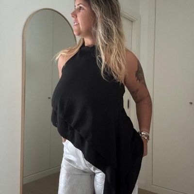 Pessoa com top preto assimétrico e calças brancas em ambiente interior com espelho e porta