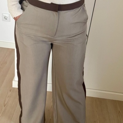 Calças bege com risca lateral castanha e cinto castanho, usadas com blusa bege, em ambiente interior com chão de madeira.