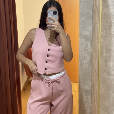 Mulher com colete e calças rosa numa divisão com porta de madeira e parede cinza