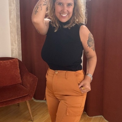 Mulher vestindo calças cargo ferrugem e top preto num interior com cadeira castanha