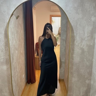 Mulher a experimentar vestido preto longo em espelho oval num provador com cortina castanha.