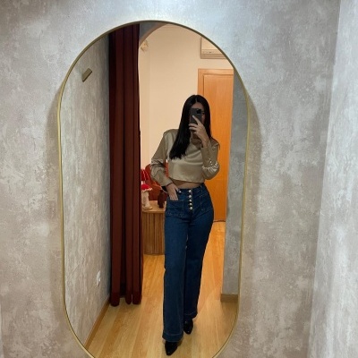 Espelho oval com moldura dourada refletindo mulher com blusa bege e calças de ganga