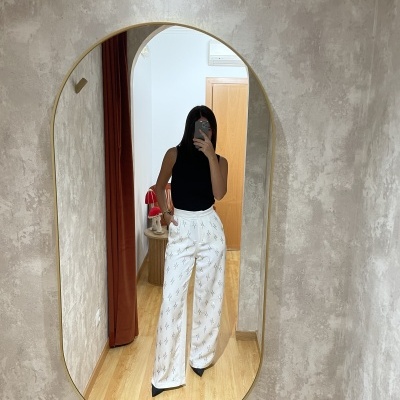 Calças largas brancas com padrão de texto e top preto refletidos em espelho oval com moldura dourada num interior de parede cinzenta texturizada