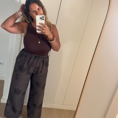 Mulher de top castanho e calças largas cinzentas com padrão de flores a tirar selfie no espelho