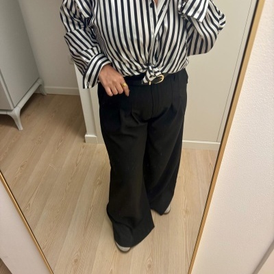 Pessoa com camisa de riscas pretas e brancas e calças pretas largas, tirando foto ao espelho num quarto com chão de madeira