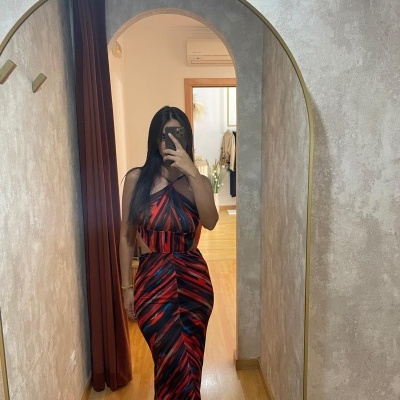 Vestido longo listrado vermelho e preto com cinto largo na cintura, refletido em espelho oval numa sala com parede cinza e cortina castanha.