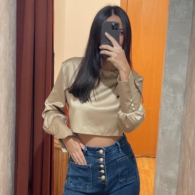 Mulher com blusa cropped bege acetinada e jeans azul com botões dourados a tirar selfie em espelho.